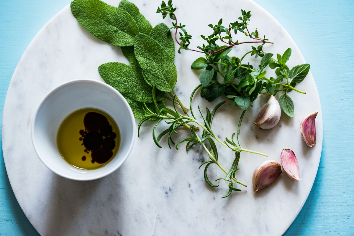 A marble surface with a bowl of olive oil and balsamic vinegar, fresh herbs, and garlic cloves