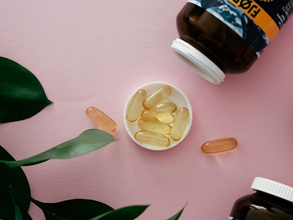 Omega 3 capsules spill from a black bottle onto a pink surface, surrounded by green leaves and plants