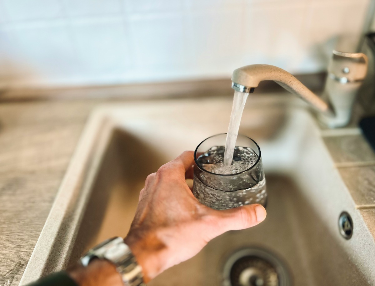 A hand holding a glass under a kitchen faucet with tap water flowing into the glass