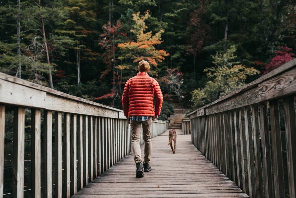 Person in a red jacket walking a dog across a wooden bridge surrounded by colorful autumn trees